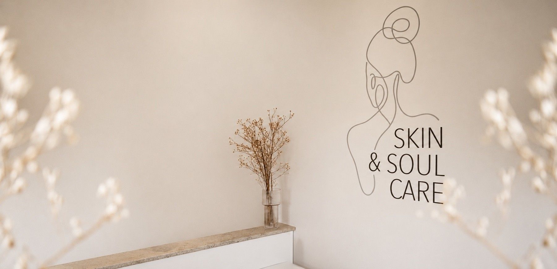 Heller Empfangsraum mit dem Logo an der Wand, davor eine Holzbank mit Trockenblumen