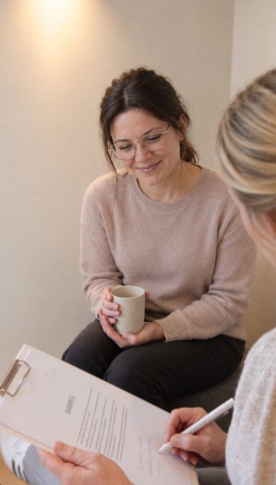 Maike notiert auf einem Klemmbrett, ihr gegenüber sitzt eine Kundin mit einer Tasse in den Händen und lächelt