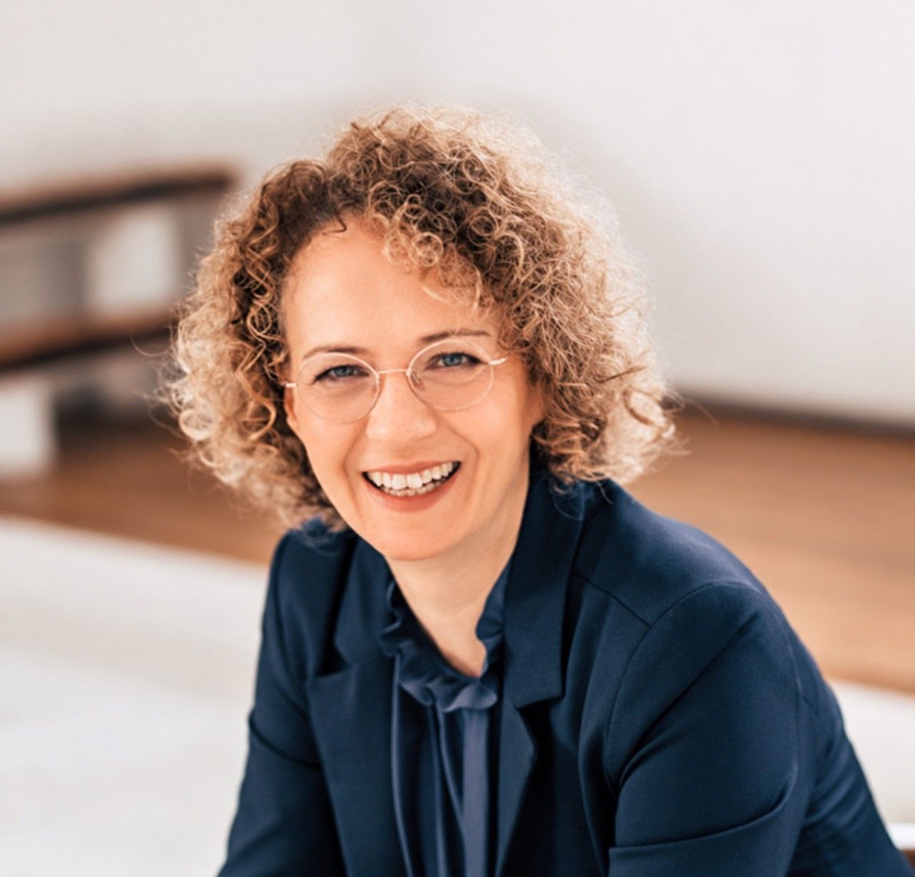 Ina Matthias mit lockigem Haar, runder Brille und dunkelblauem Blazer lächelt in die Kamera